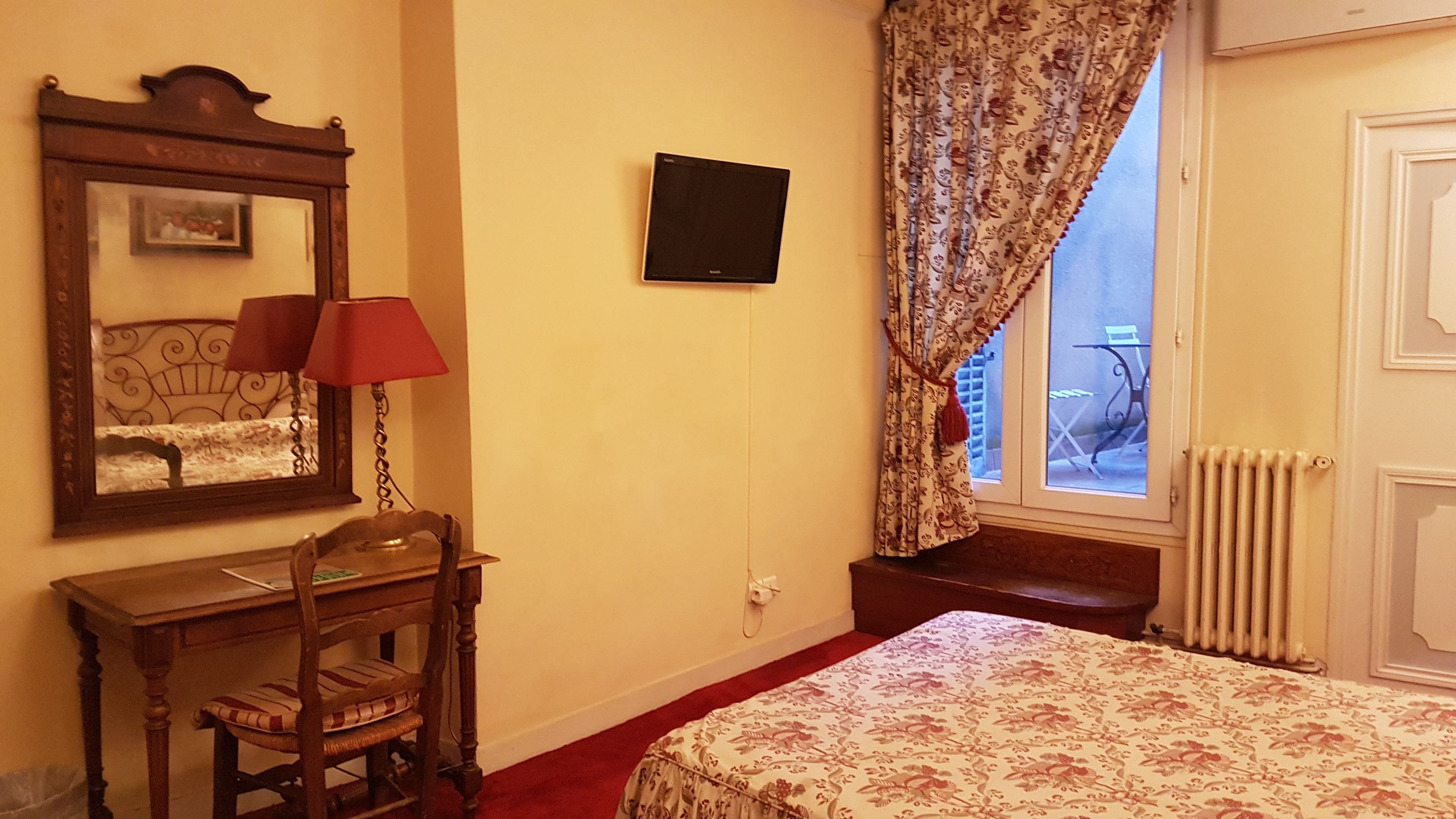 Une chambre à coucher avec un lit, un bureau avec une chaise, un miroir, une télévision et une fenêtre avec des rideaux.