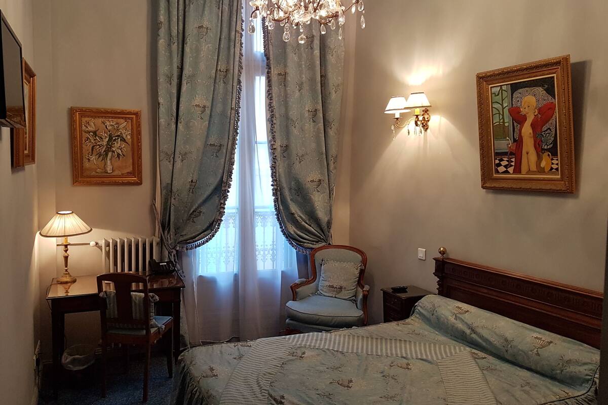 A hotel room with a bed, a chair, a desk with a lamp, a radiator, and a window with curtains.