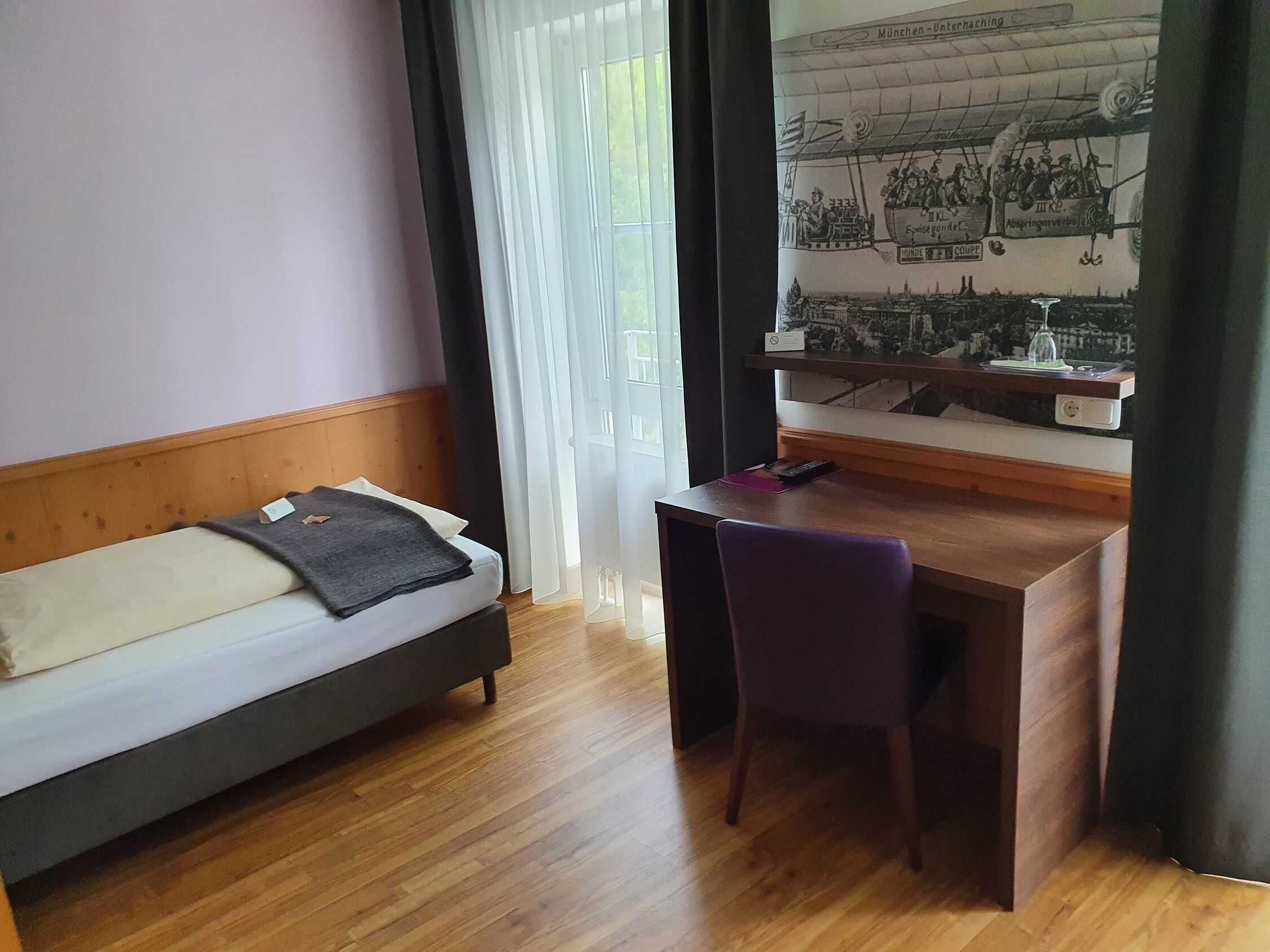 Une pièce comprenant un lit, un bureau et une chaise. Il y a une fenêtre avec des rideaux et un mur orné d’une fresque.