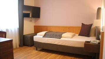 Quarto de hotel com cama, mesa de cabeceira, luminária e TV fixada na parede.