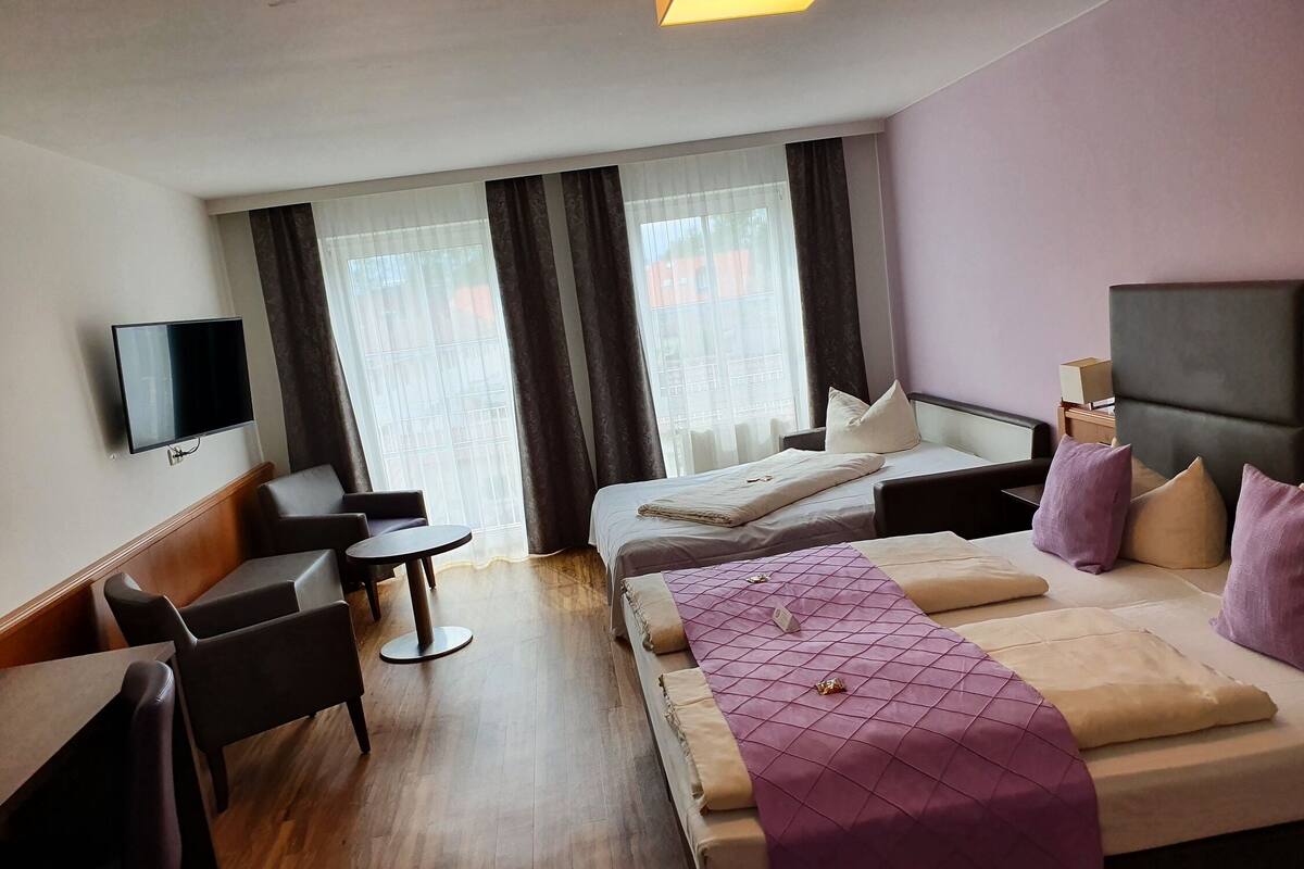 Een hotelkamer met een bed, een bureau, een stoel, een televisie en grote ramen met gordijnen.