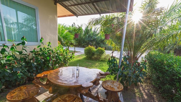A patio with a round table and four stools, surrounded by lush greenery and a house in the background. - Huasai Palm Inn (Hua Sai)