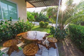A patio with a round table and four stools, surrounded by lush greenery and a house in the background. - Huasai Palm Inn (Hua Sai)