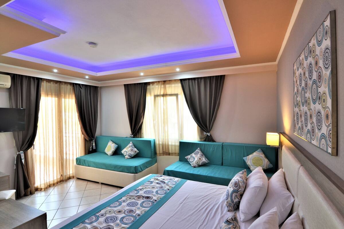 A hotel room with two green sofas, a bed with patterned bedding, and a ceiling with blue lighting.