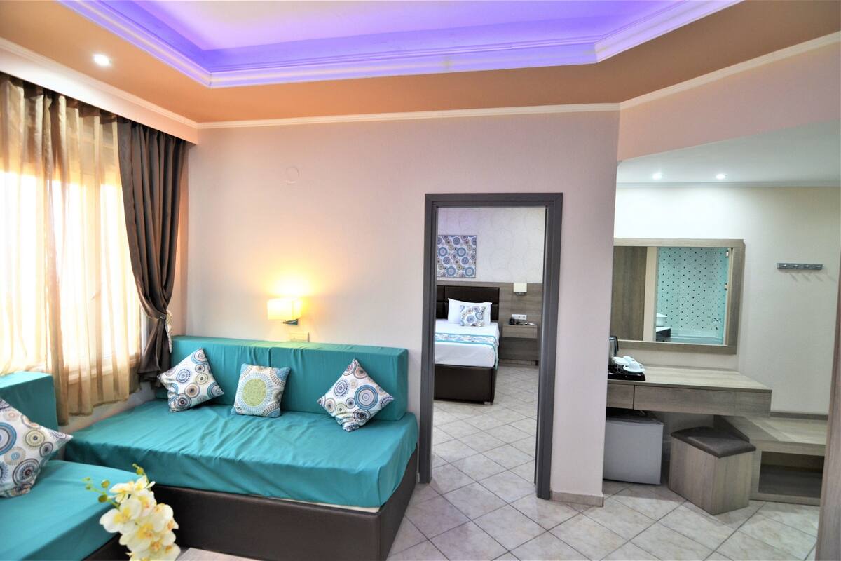 A hotel room with a teal sofa, a bed with patterned bedding, a small desk with a mirror, and a bathroom with a toilet and sink.