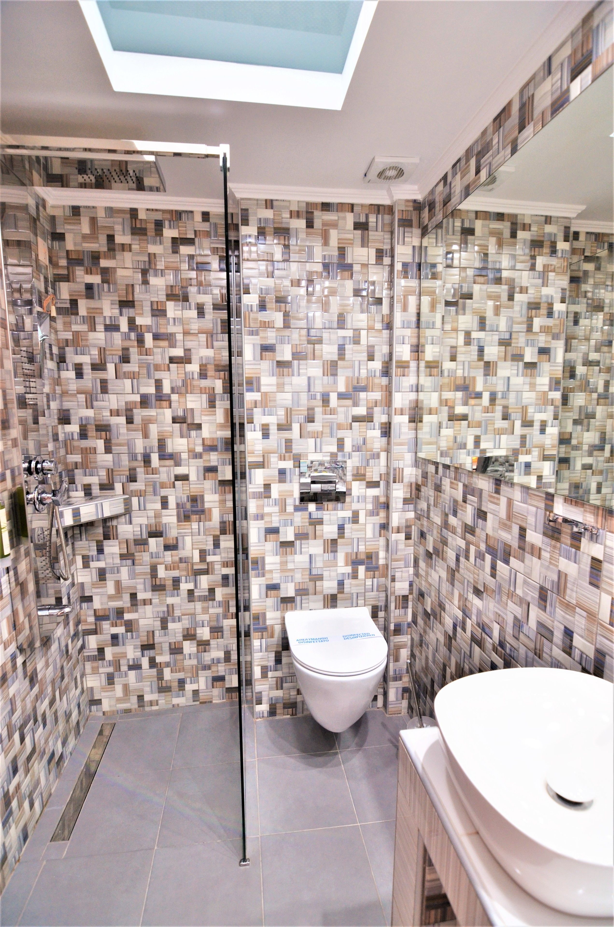 A modern bathroom with a mosaic tile wall, a skylight, a shower, and a white toilet.