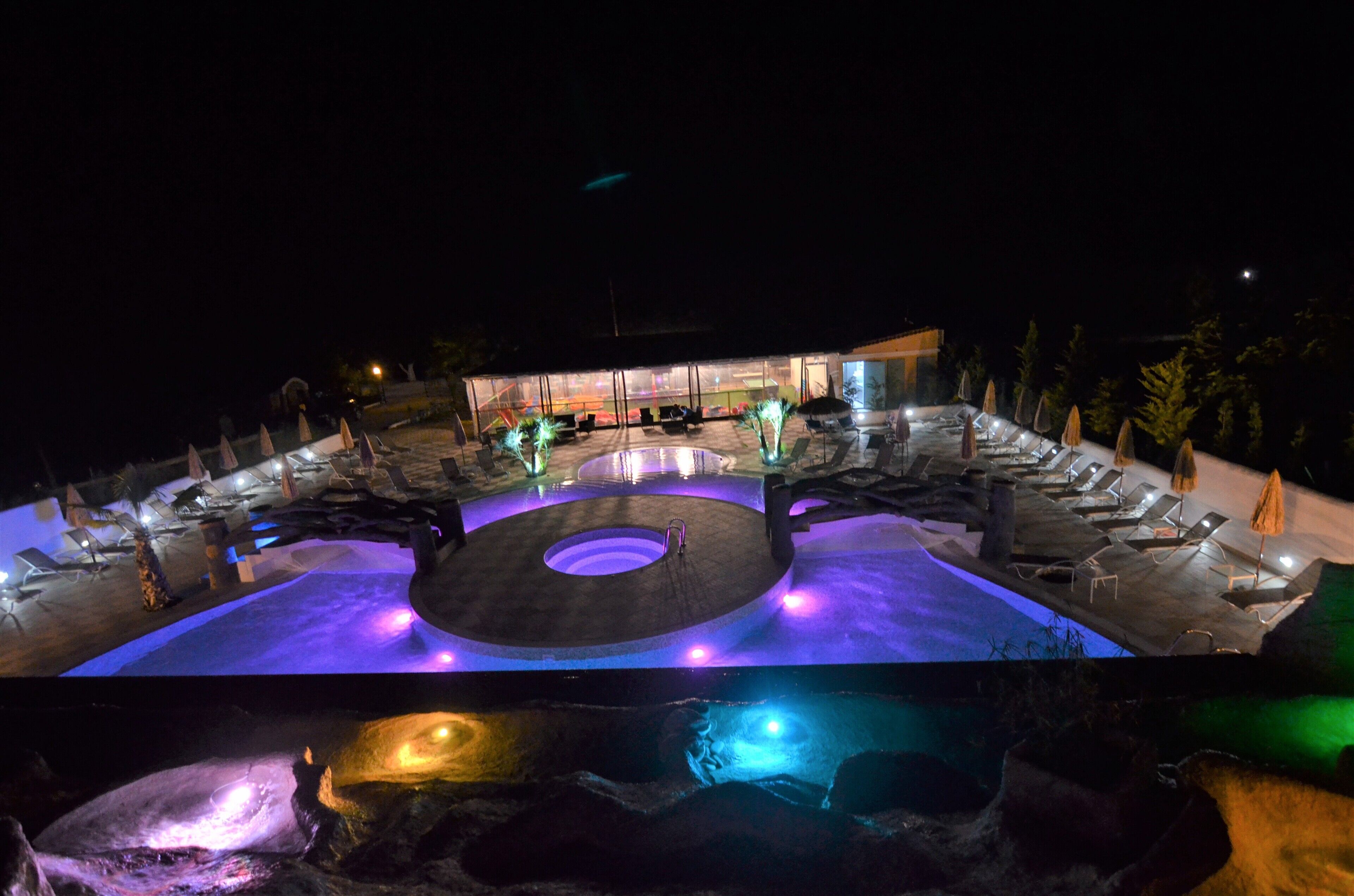 Ein Nachtblick auf einen Hotelpoolbereich mit mehreren Liegestühlen und Sonnenschirmen, einer zentralen runden Wasserspiels und einem Gebäude im Hintergrund.
