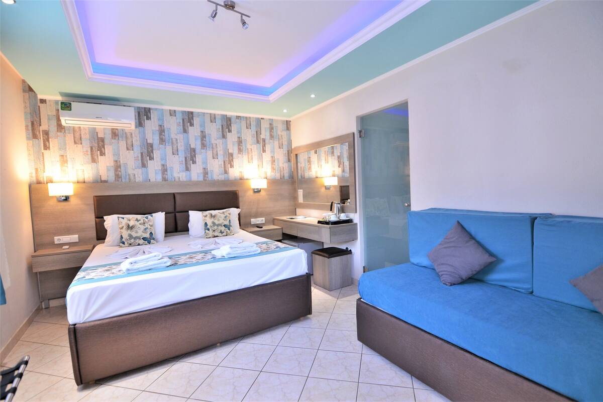 A modern hotel room with a large bed, a sofa, a vanity with a mirror, and a wall-mounted air conditioner.