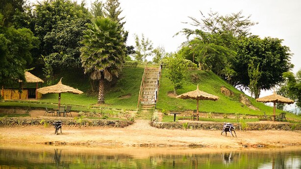 A serene lakeside setting with a stone staircase leading up a grassy hill, surrounded by trees and thatched-roof structures.