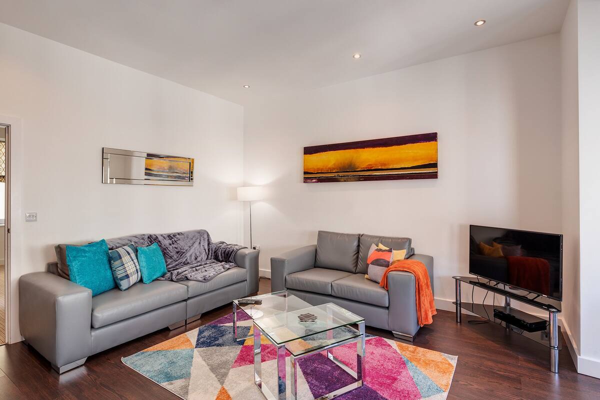 A modern living room with two grey sofas, a glass coffee table, a flat-screen TV on a stand, and a colorful rug.