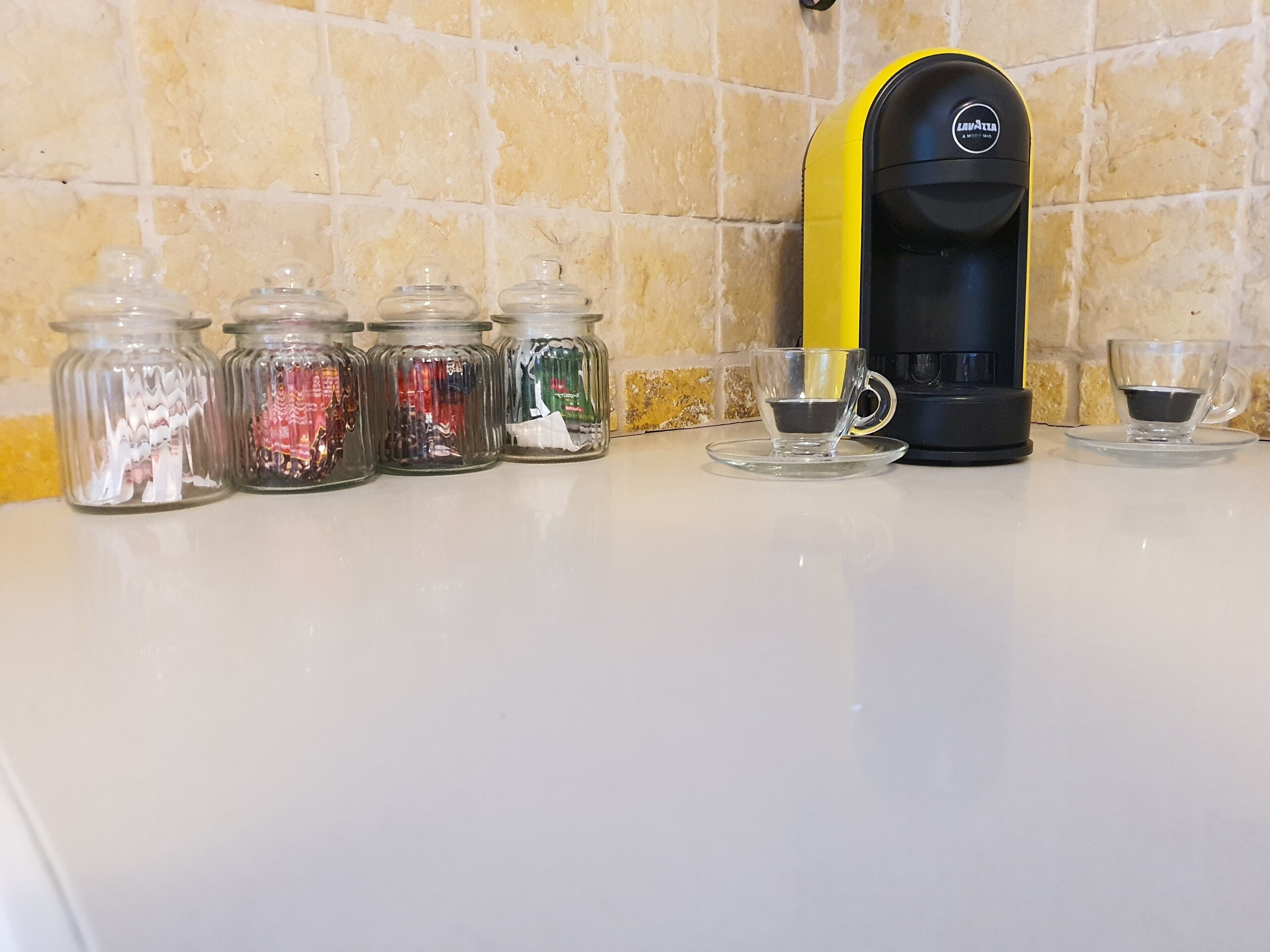 A kitchen counter with a coffee maker, four glass jars with various items, and two empty cups.