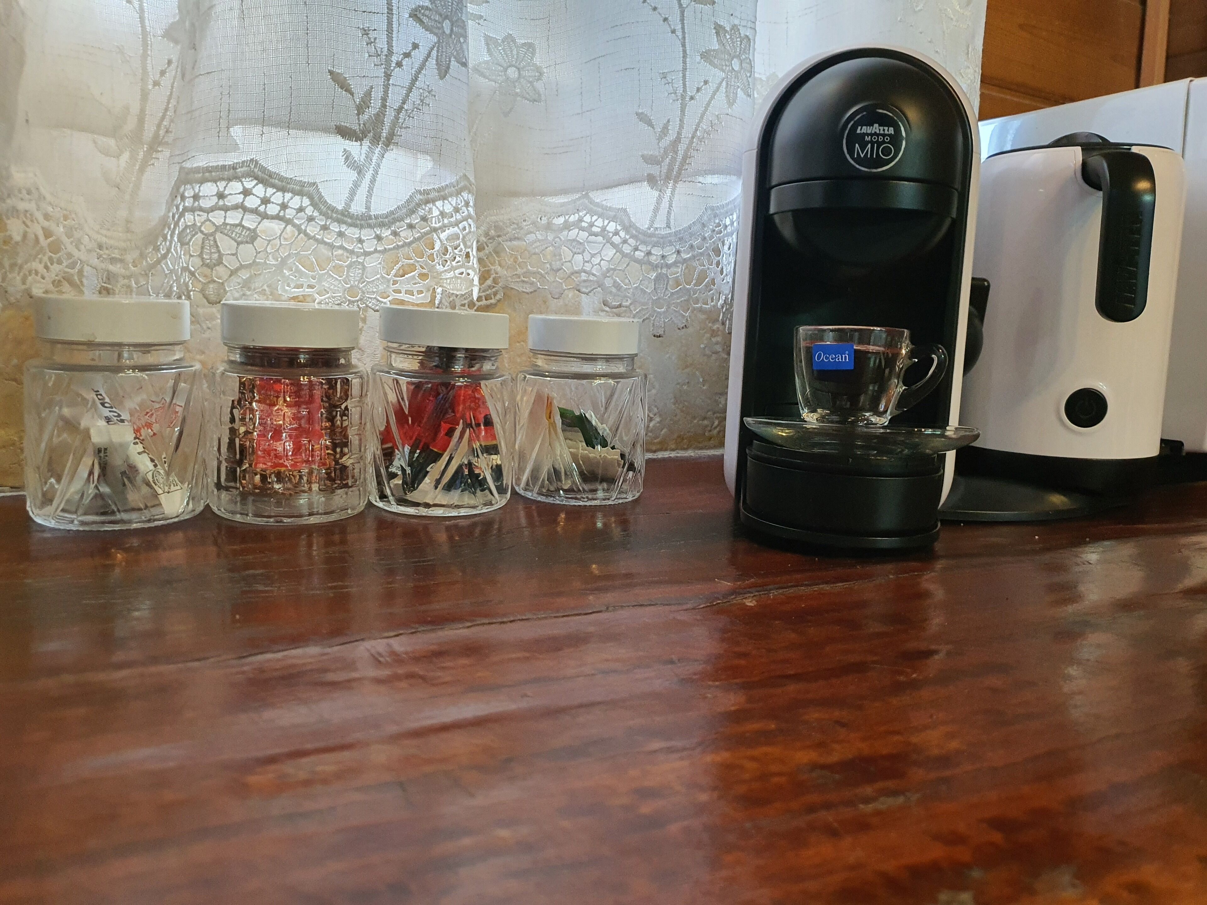 Una encimera de cocina con una cafetera, cuatro frascos de especias y una cortina de encaje al fondo.