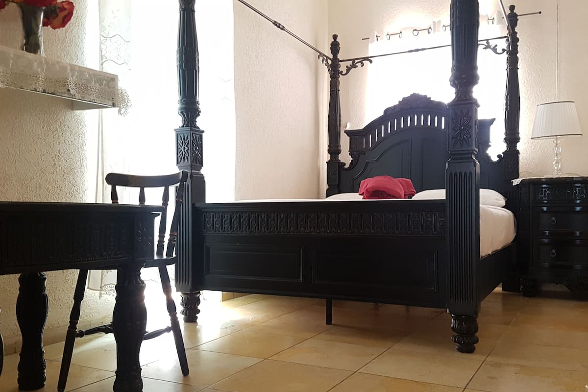 Un lit à baldaquin avec une finition sombre, une chaise en bois, une petite table, une table de chevet et une lampe.