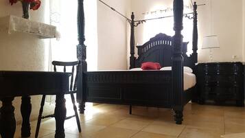 Uma cama de dossel com acabamento escuro, uma cadeira de madeira, uma mesinha, um criado-mudo e um abajur.