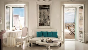 A room with a white sofa, teal cushions, a coffee table with books, and a view through an open door to the outside. - B&B Centro Storico (Otranto)