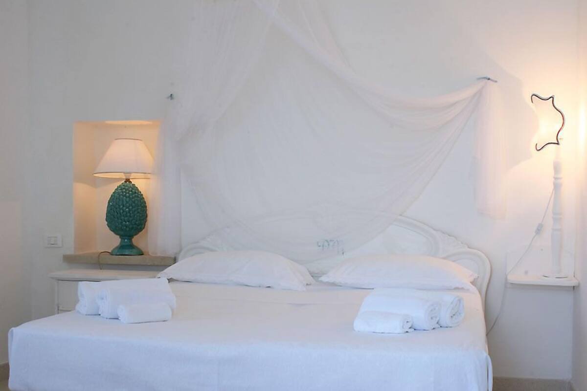 Una cama bien hecha con sábanas y toallas blancas, una lámpara verde sobre una mesita de noche y una lámpara de pared.