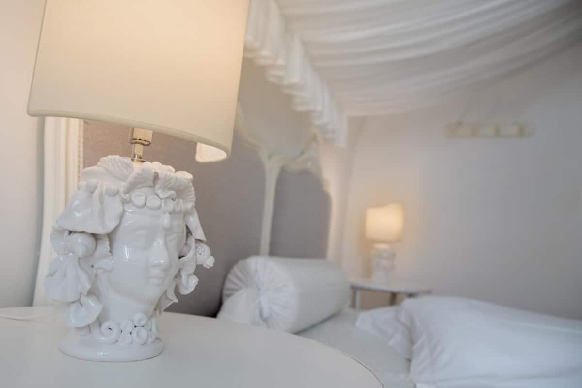 Una habitación blanca con un jarrón decorativo blanco sobre una mesa, una cama con ropa de cama blanca y una lámpara blanca.