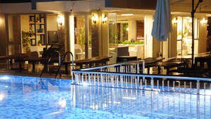Une piscine intérieure avec un sol carrelé, entourée de chaises longues et de tables, et une paroi en verre qui sépare l’espace piscine de l’espace salon adjacent.