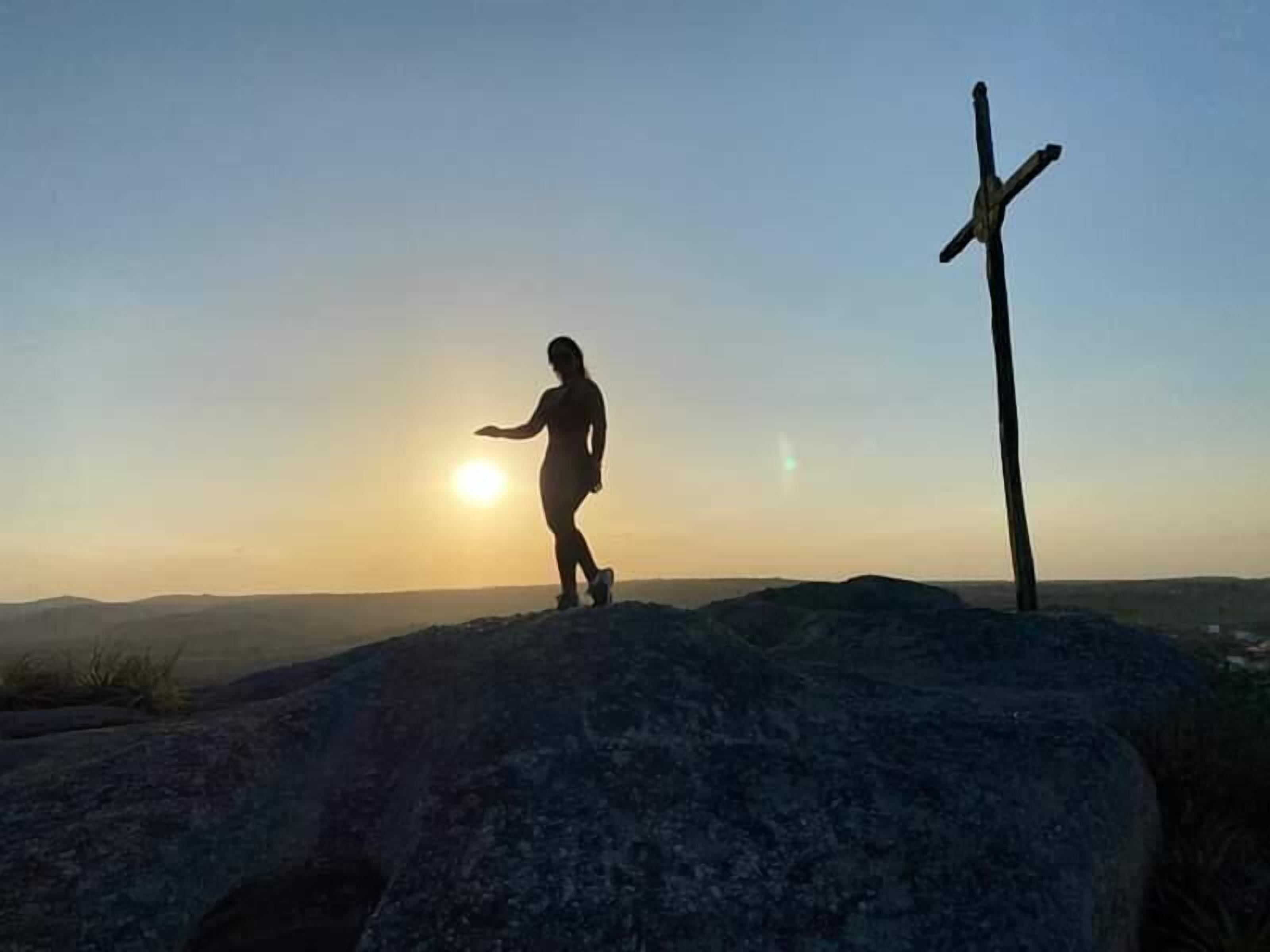 Una persona de pie sobre una roca con una cruz al fondo durante el atardecer.