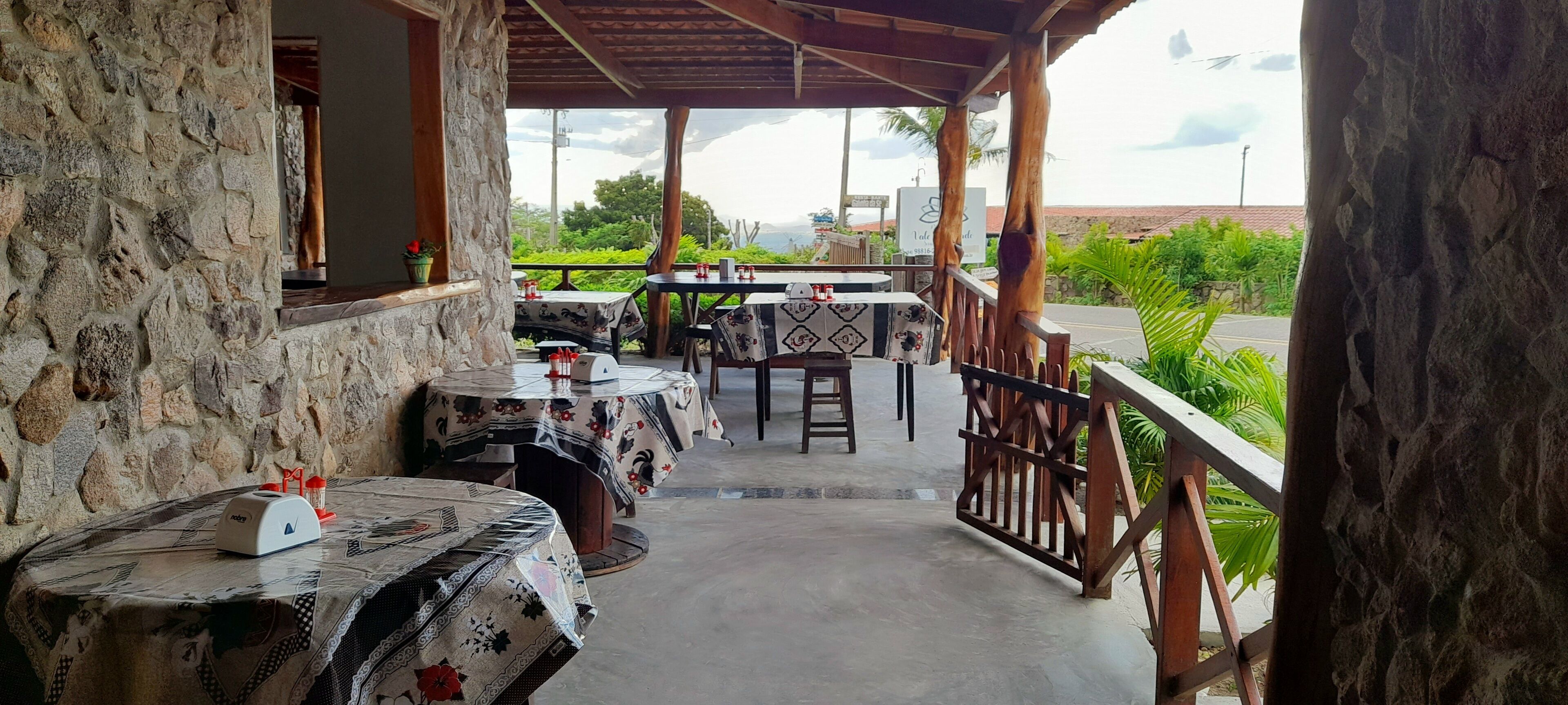 Un área de comedor al aire libre cubierta, con paredes de piedra, mesas con manteles estampados y vistas a zonas verdes y un paisaje lejano.