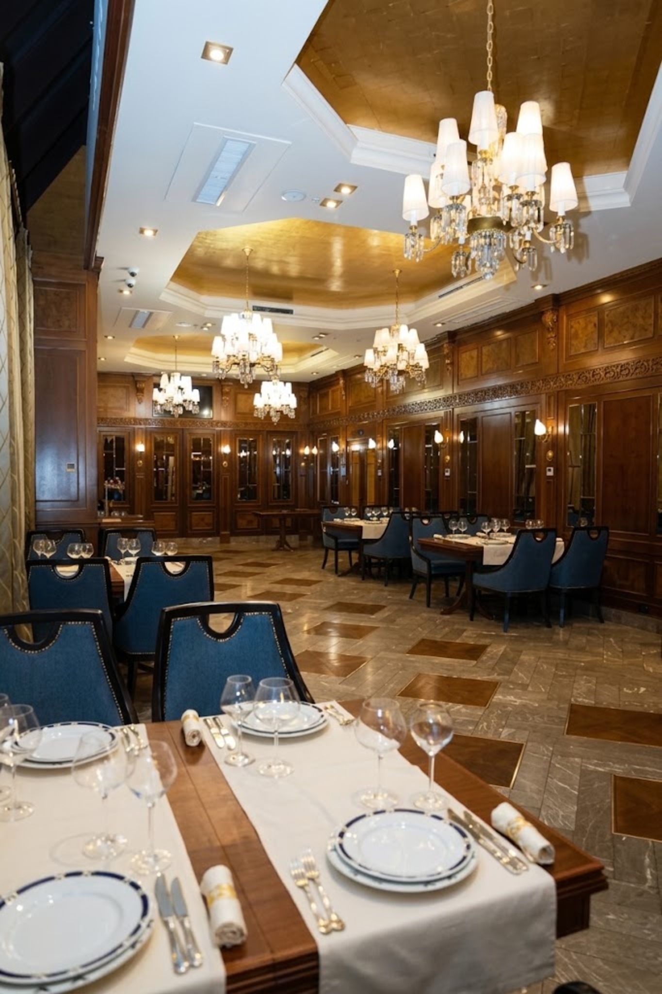 A formal dining area with multiple tables set for service, elegant chandeliers, and a classic interior design.