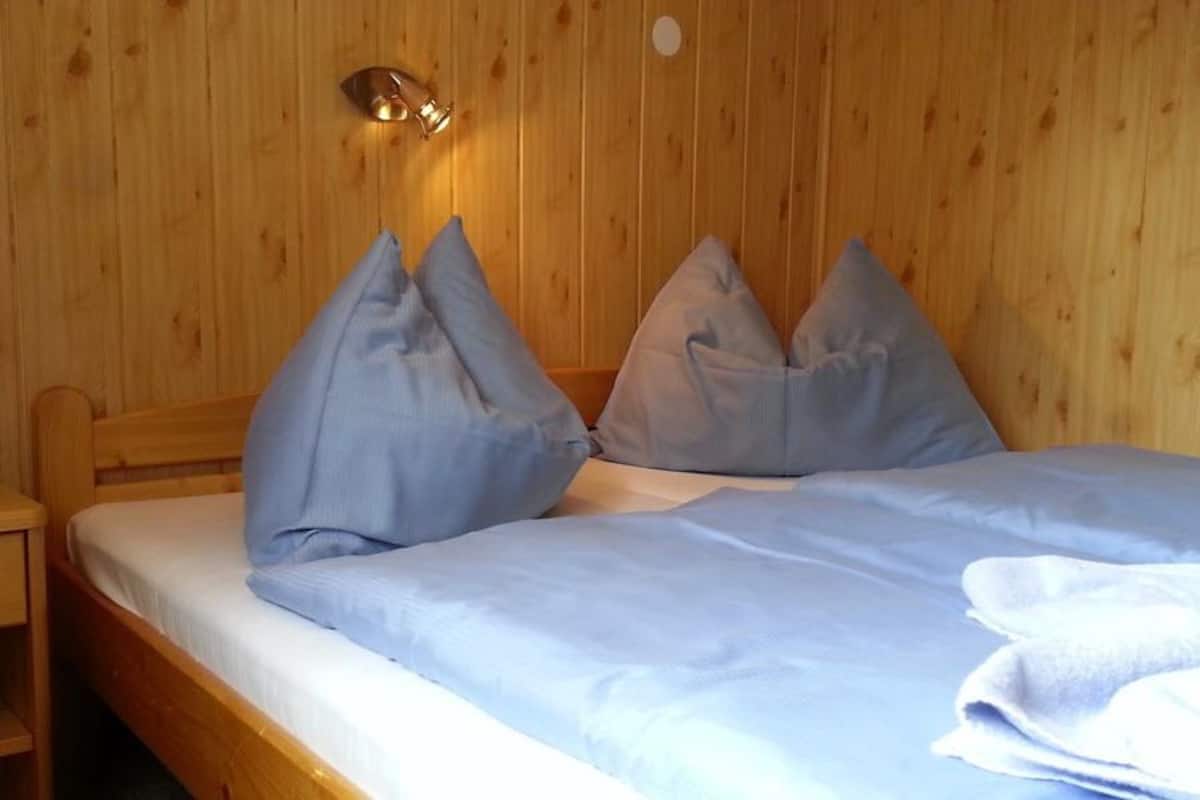 Quarto com painéis de madeira, uma cama, duas almofadas, uma mesinha de cabeceira e um aplique na parede.