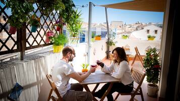 A couple seated at a wooden table on a balcony with potted plants and a city view.