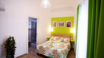 A bedroom with a bed, a potted plant, a bedside table, and a wall with three pictures.
