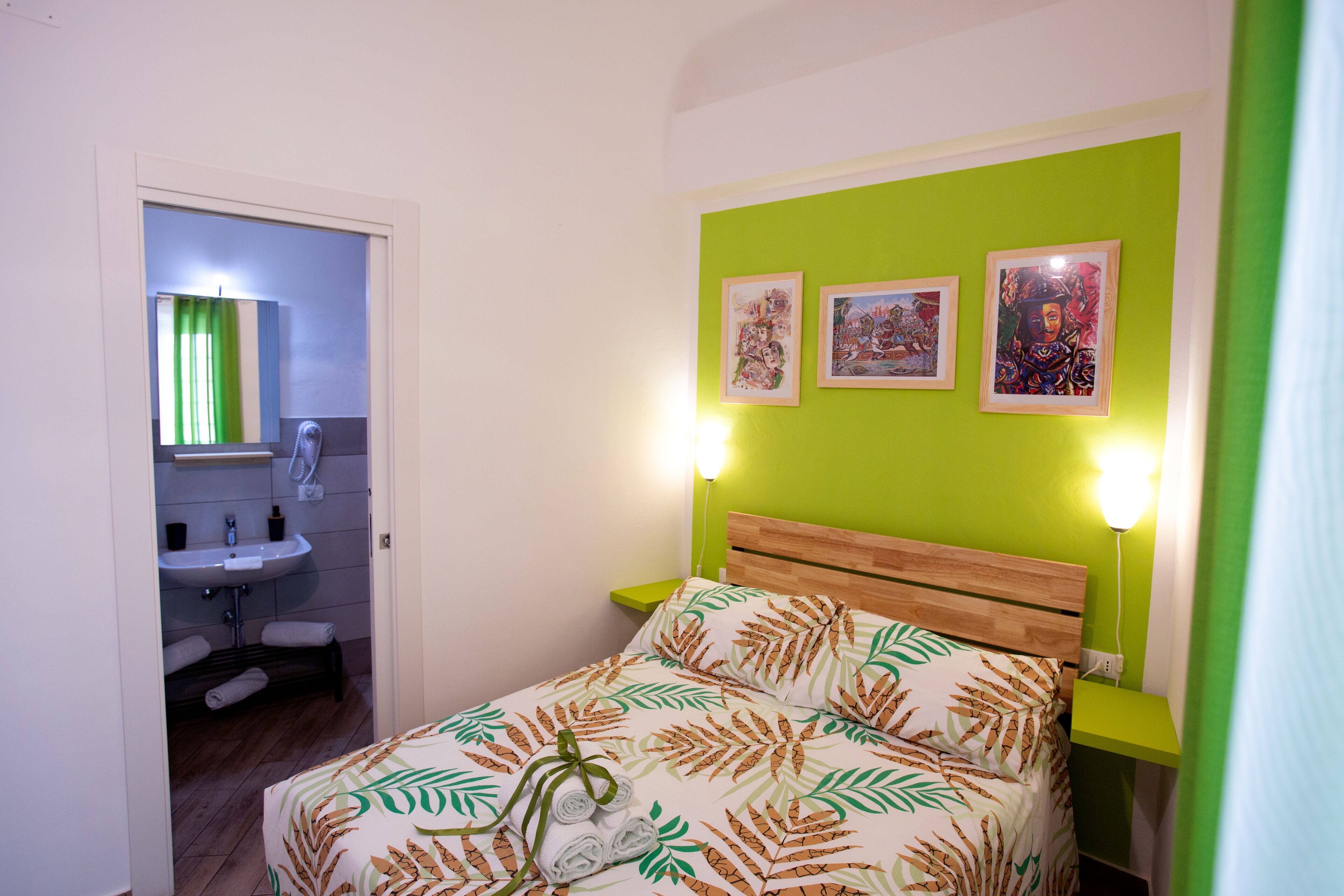 Un dormitorio con cama, pared verde, cabecero de madera, mesita de noche, lámpara y baño con lavabo y toallero.