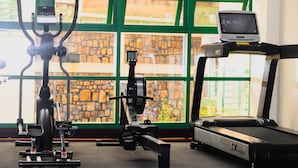 A gym with exercise equipment, including a treadmill, stationary bikes, and a rowing machine, with large windows and a stone wall in the background. - Highlands Suites Hotel (Kigali)