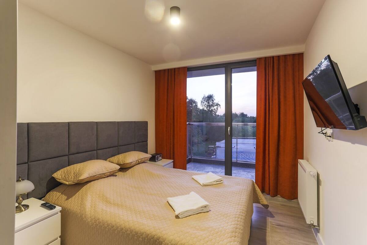 Ein modernes Hotelzimmer mit einem großen Bett, einem flachbildfernseher an der Wand und einem Balkon mit orangefarbenen Vorhängen.