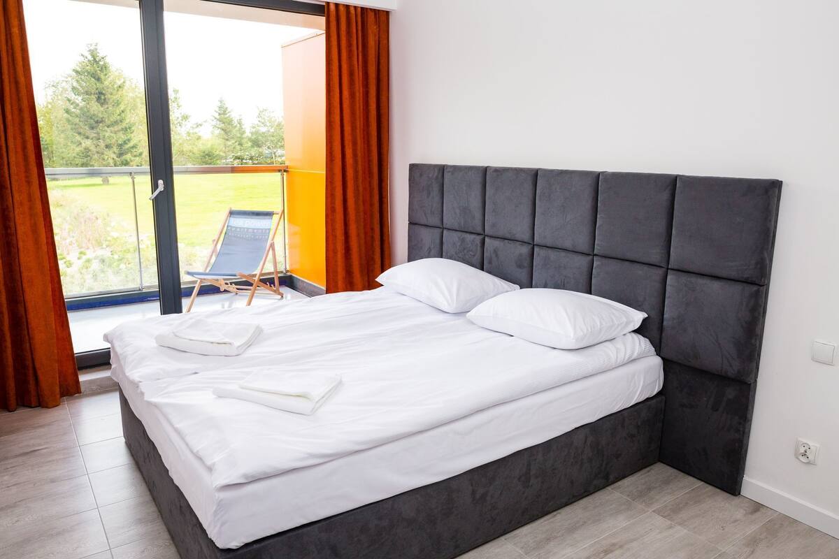 Ein ordentlich bezogenes Bett mit weißen Leinen und Kissen, ein dunkelgrau gepolsterter Kopfteil und Blick auf einen Balkon mit Stuhl und Vorhängen.