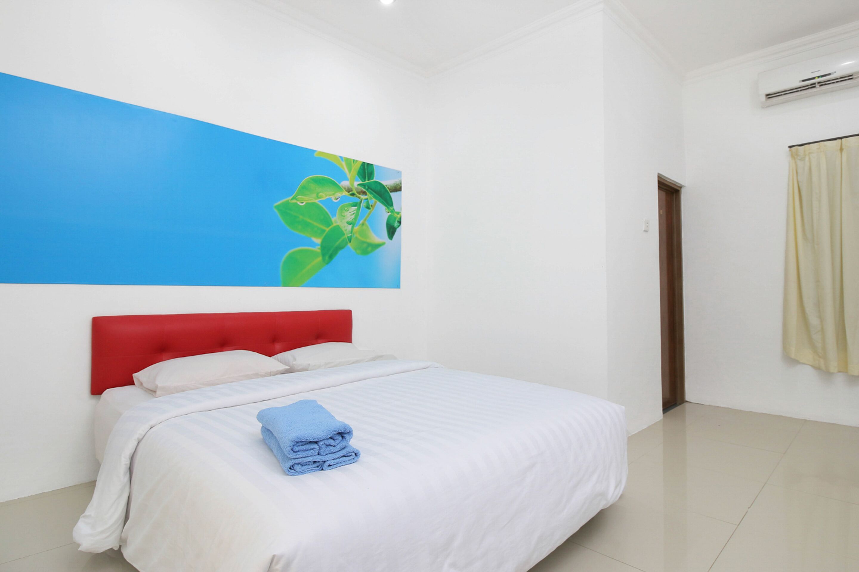 Habitación de hotel con cama, ropa de cama blanca, cabecero rojo, obra de arte azul en la pared y ventana con vista a follaje.