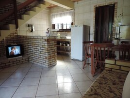 Cozinha com parede de tijolos, geladeira branca e área de refeições de madeira com uma TV à esquerda.