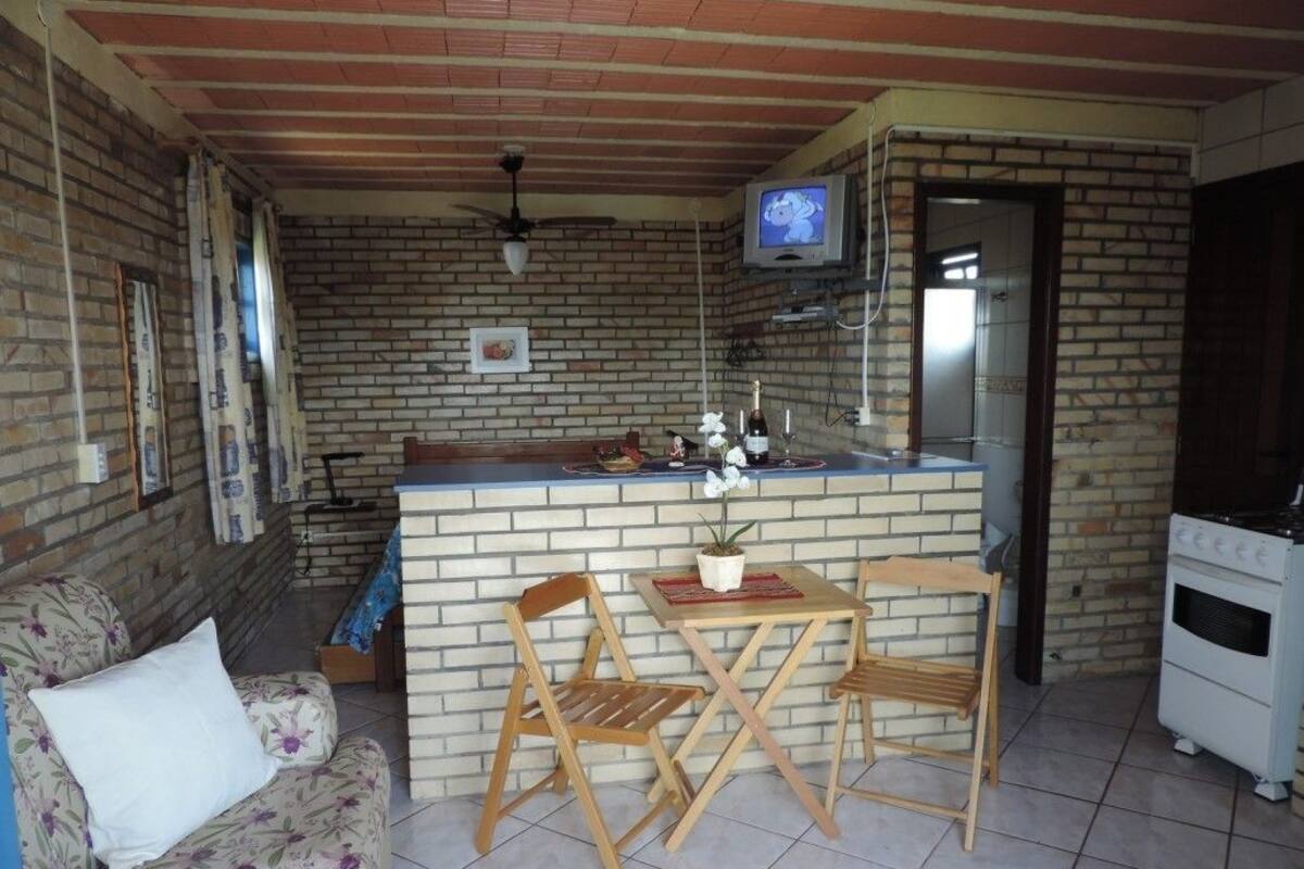Un espace de vie compact comprenant un coin cuisine, une petite table à manger, deux chaises en bois, un canapé à motifs floraux, un téléviseur fixé au mur et un réfrigérateur.