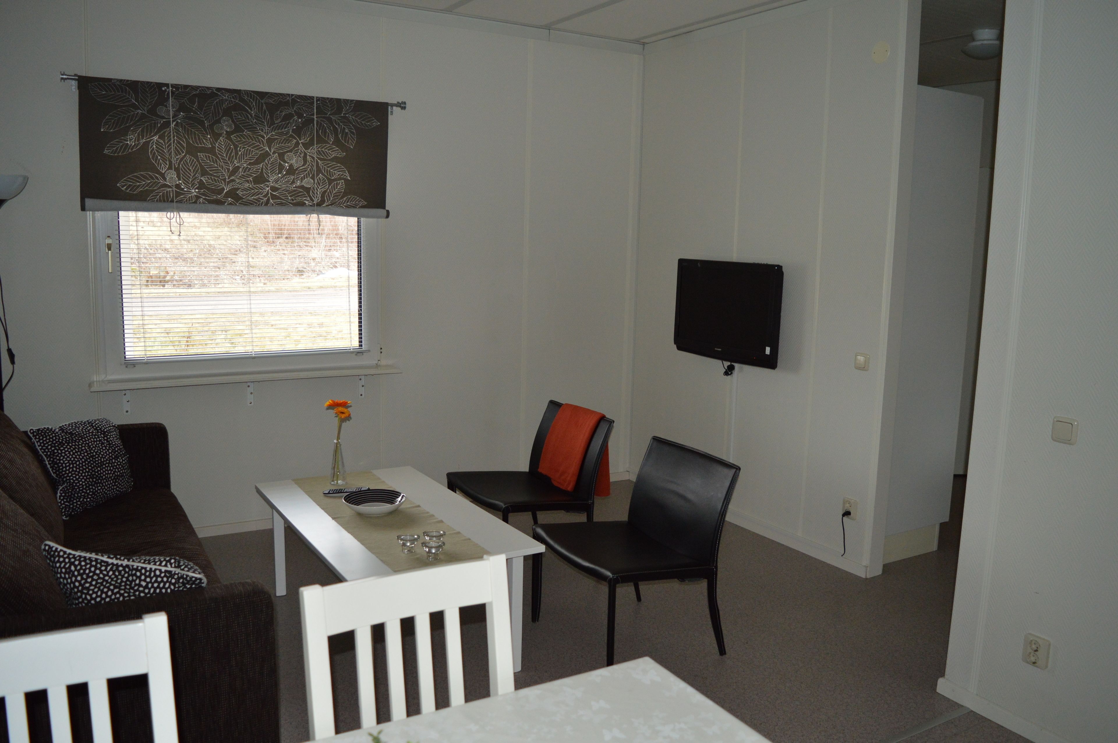 A small, sparsely furnished room with a single window, a flat-screen TV mounted on the wall, a white table, and two chairs.