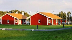Deux bâtiments rouges aux contours blancs et aux toits orange, probablement des granges ou des entrepôts, se dressent sur un champ verdoyant, avec des drapeaux en arrière-plan.
