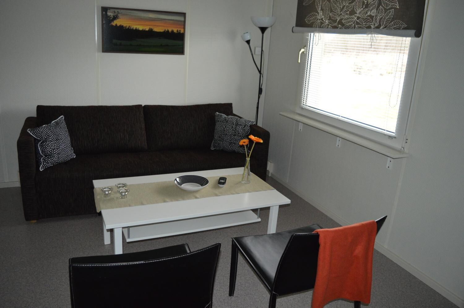 Una sala de estar con un sofá marrón oscuro, una mesa de centro blanca, una silla negra y una ventana con persianas.
