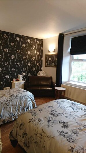 A room with two beds, a chair, a small table, a radiator, and a window with curtains.