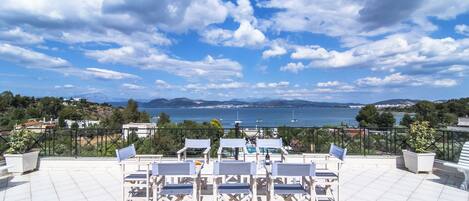 A terrace with white chairs and tables, overlooking a scenic view of the sea and mountains.