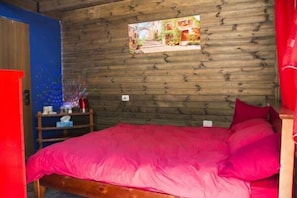 Une pièce avec un mur en bois, un lit avec une literie rose, une petite table avec une lampe rouge et un tableau encadré au mur.