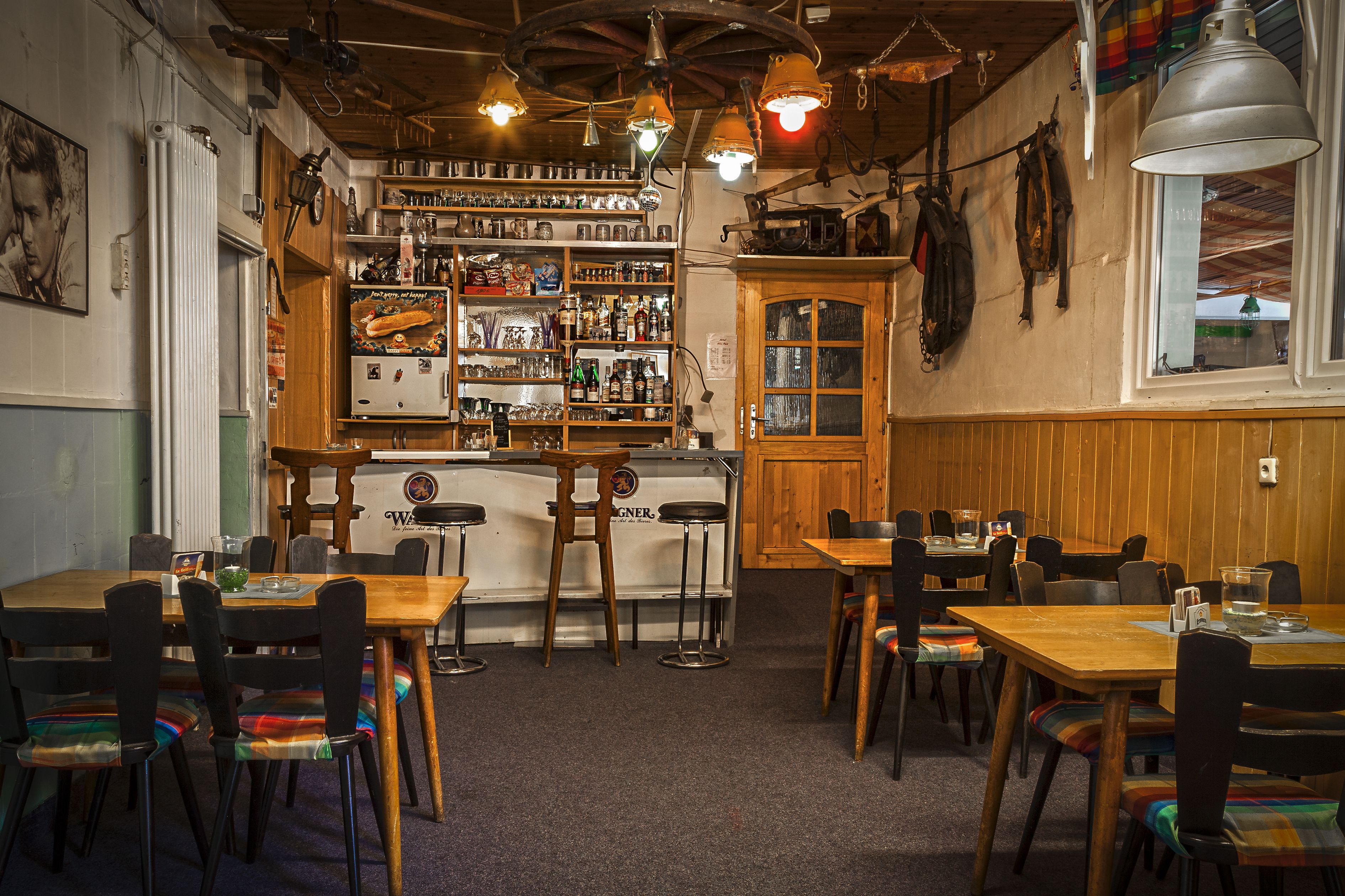 A restaurant with wooden tables and chairs, a bar area with shelves stocked with bottles, and a view through a window.