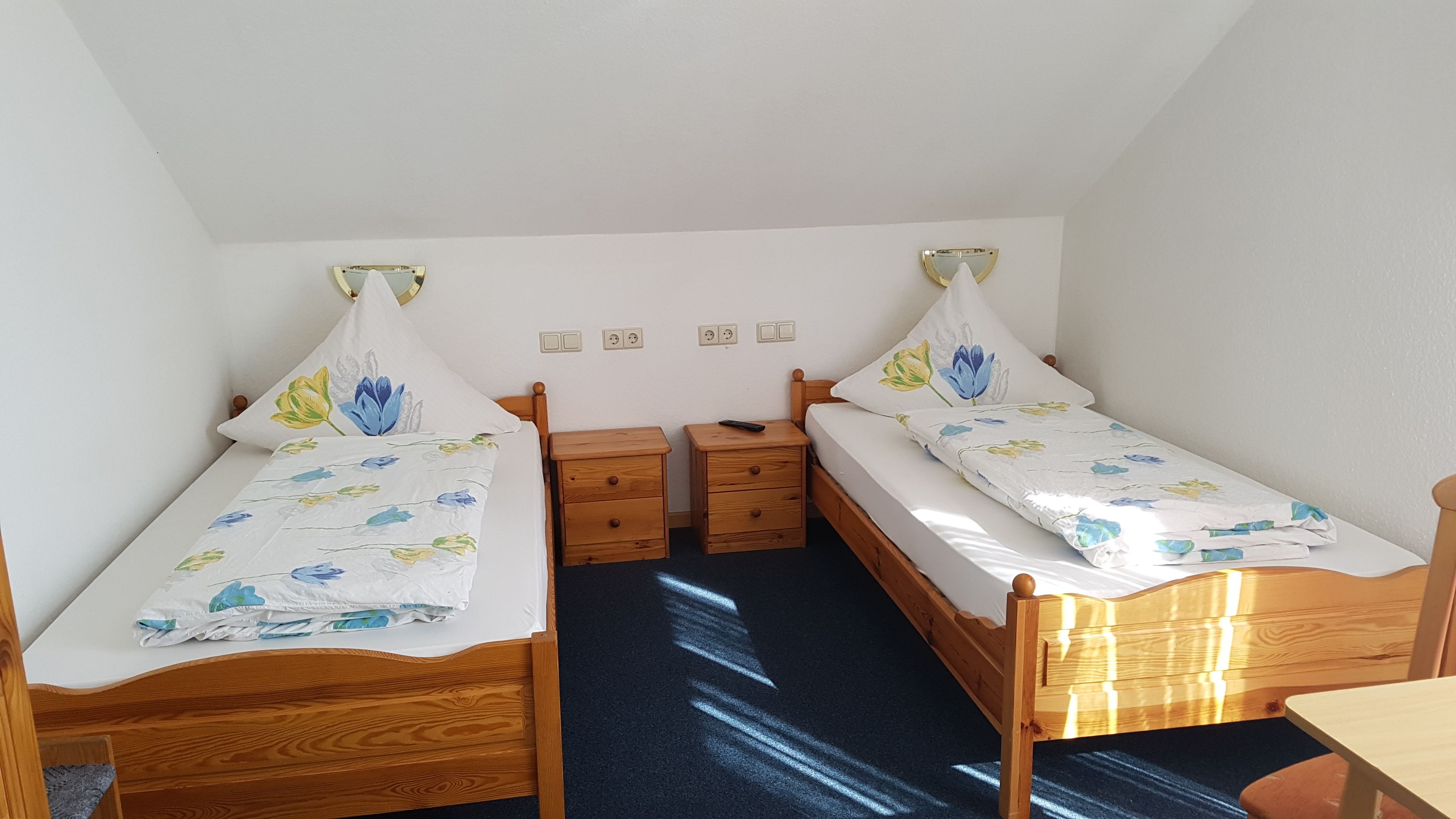 A small, clean room with two wooden beds, each with a floral bedspread and matching pillows. There are two bedside tables with drawers, a small table in front of the beds, and a wall-mounted light fixture.