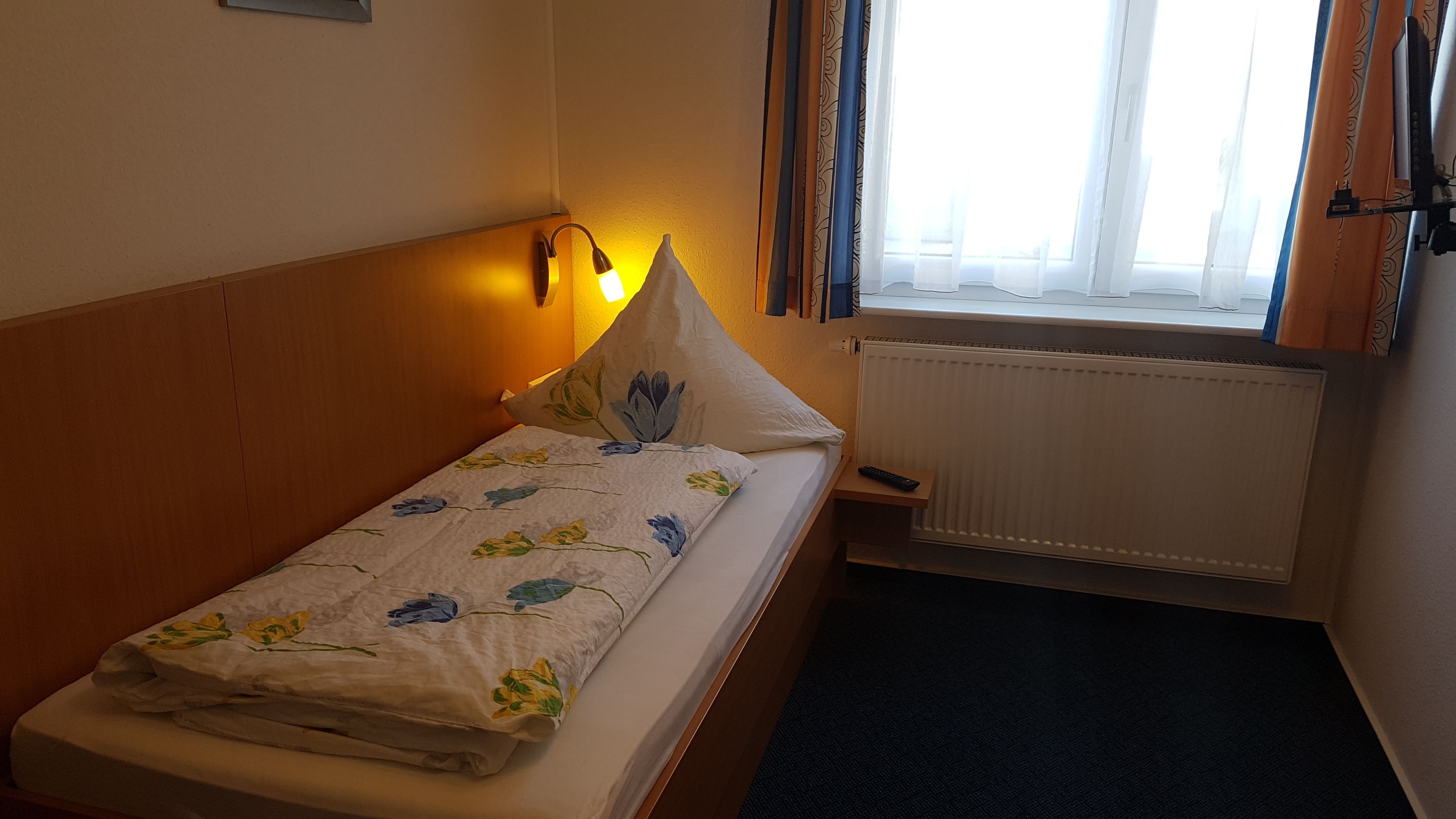 Une petite chambre avec un lit simple, une tête de lit en bois, une table de chevet avec une lampe, une fenêtre avec des rideaux et un radiateur.