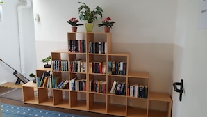A multi-tiered wooden bookshelf filled with books, topped with potted plants, against a white wall. - Hotel Alte Post (Kehl)