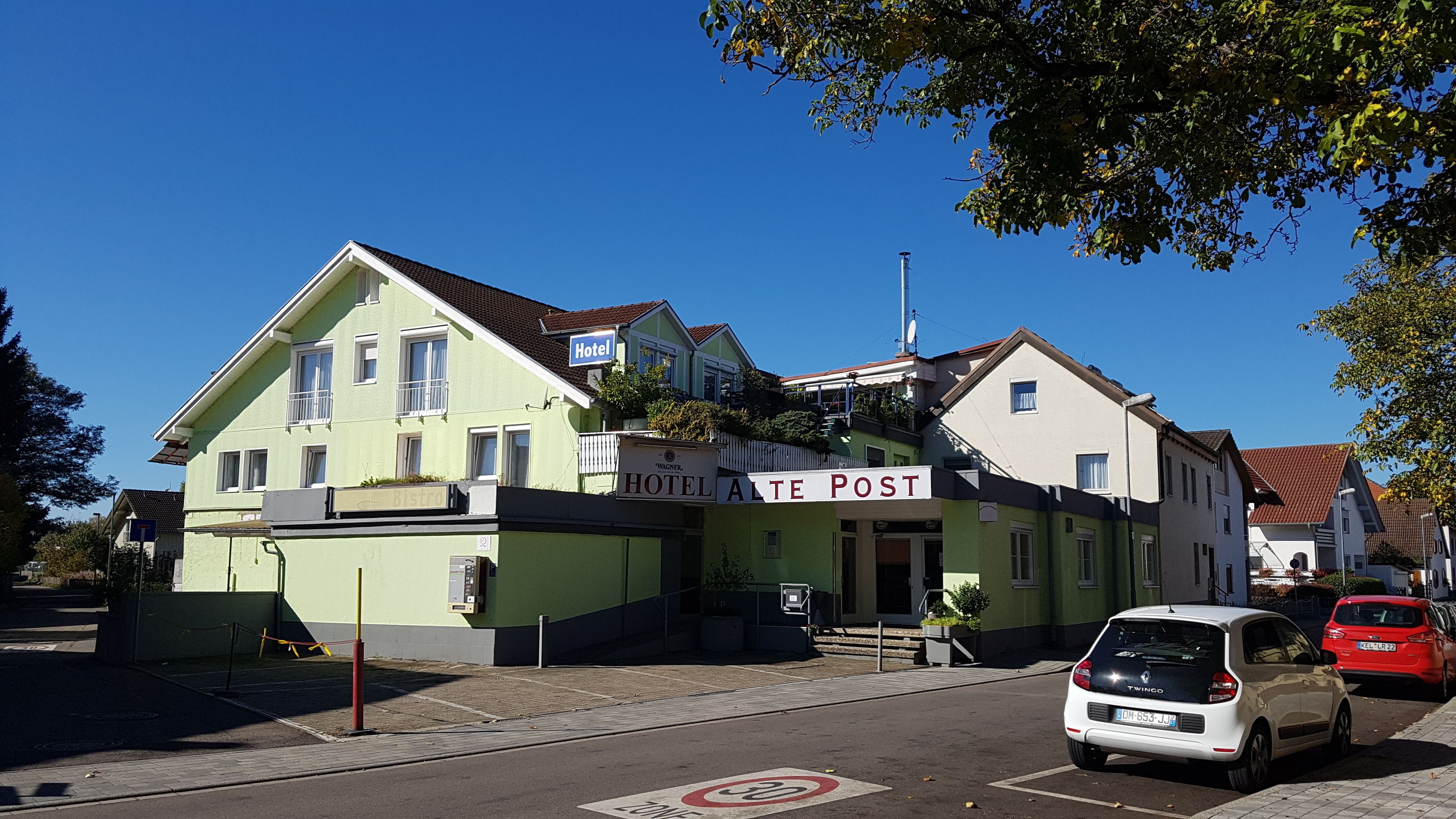 A hotel with a sign reading "HOTEL ALTE POST" and a "BISTRO" sign below it. There are cars parked in front, and a speed limit sign of 30 is visible on the road.