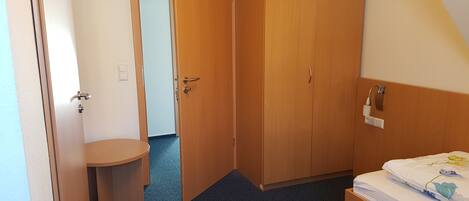 Habitación de hotel con armario de madera, una mesita de noche pequeña, cama con edredón estampado floral y una lámpara de pared.