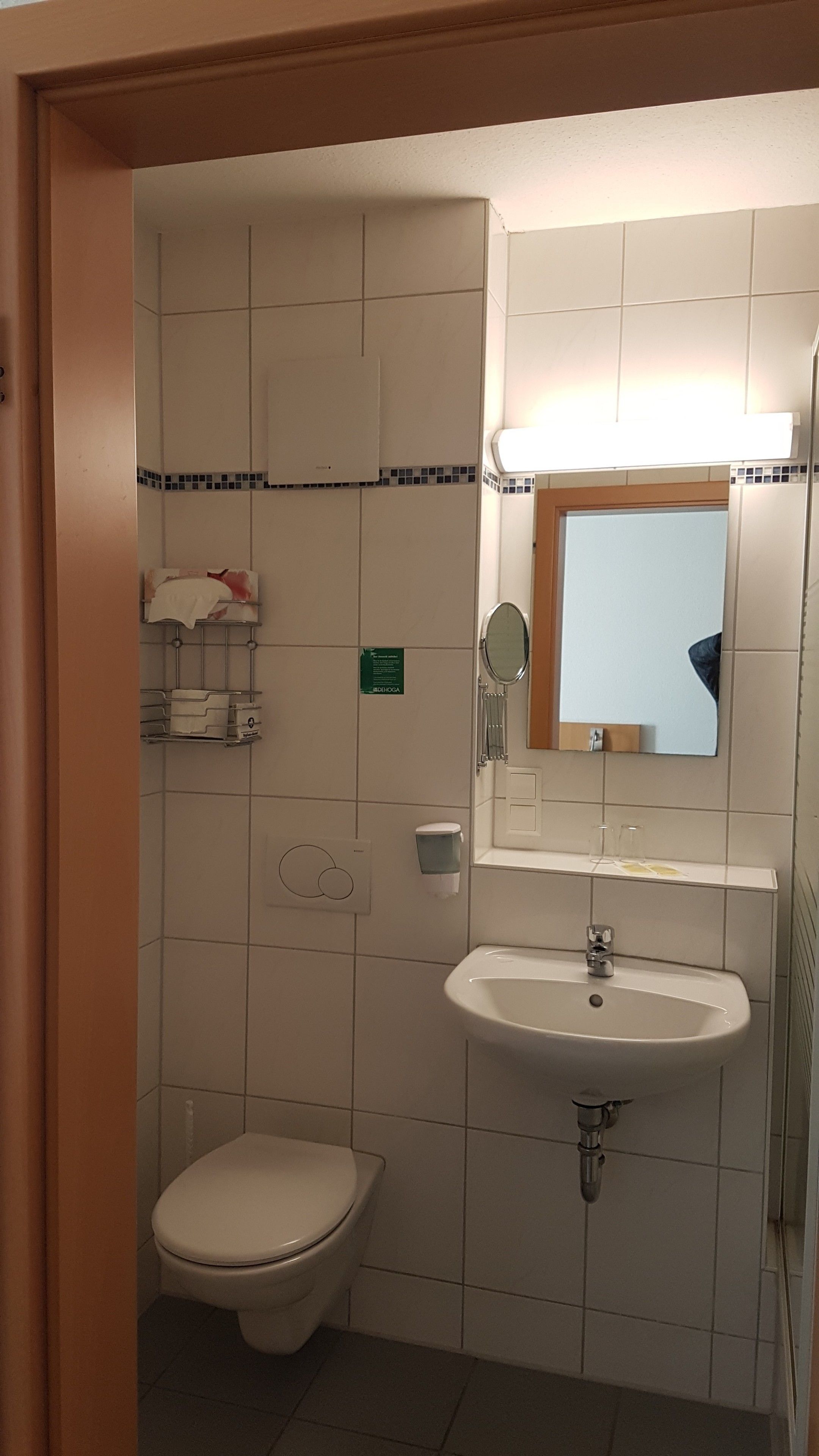 Un baño pequeño y limpio con inodoro, lavamanos y espejo. Hay un perchero sobre el inodoro y un cartel en la pared.
