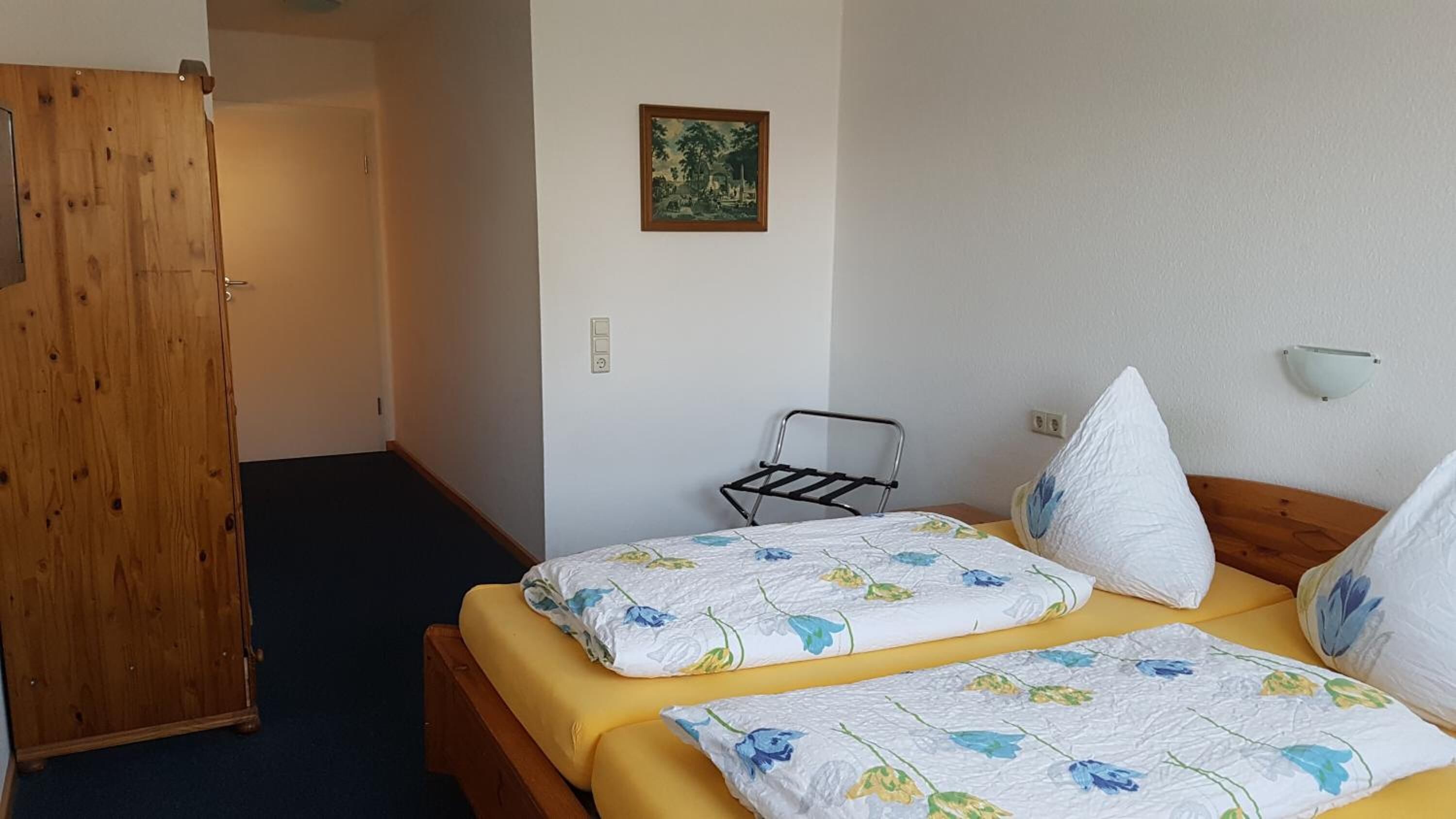 A room with two beds, a wooden wardrobe, a small table, a wall-mounted light, and a painting.