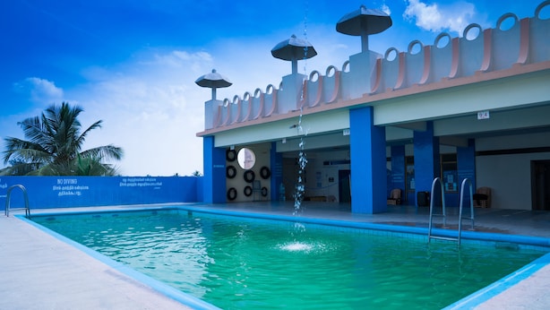 Una piscina con un cartel de prohibido bucear, un edificio con pilares azules y un cielo despejado.